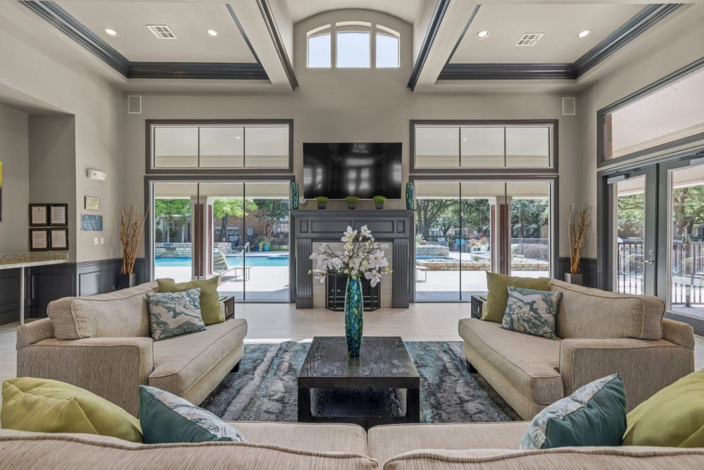 Spacious and modern clubhouse with high ceilings, large windows looking out to a pool area, two beige sofas with green and beige pillows, a central black coffee table, and a mounted flat-screen TV above a dark wood entertainment console at Brooks on Preston in Plano, TX.