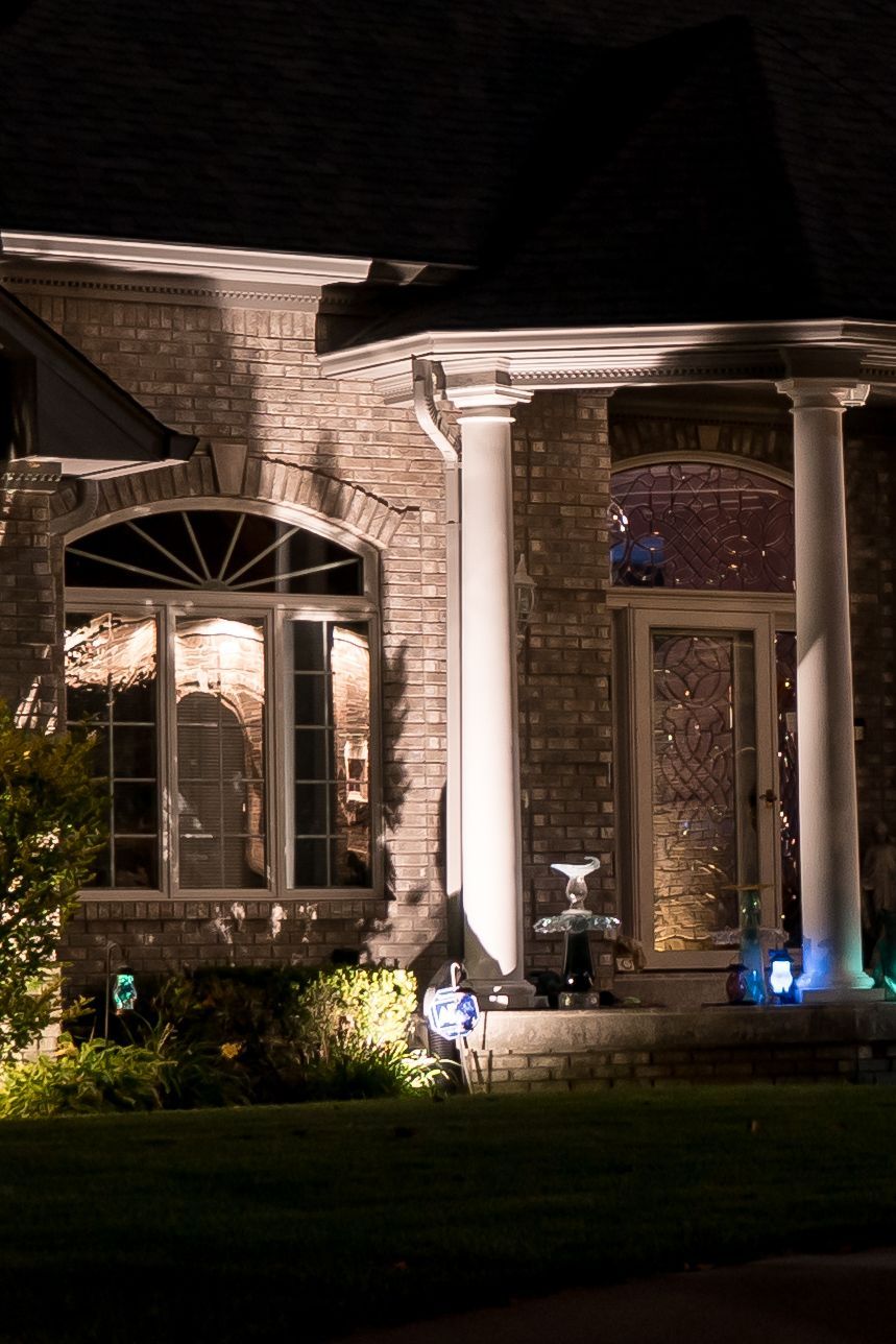 Brick house facade at night, lit by spotlights. White columns frame the front entrance.
