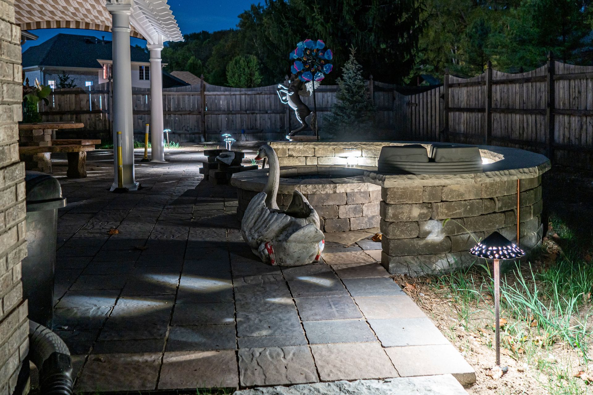 Nighttime patio with stone pavers, seating, and decorative lighting. Wooden fence in background.