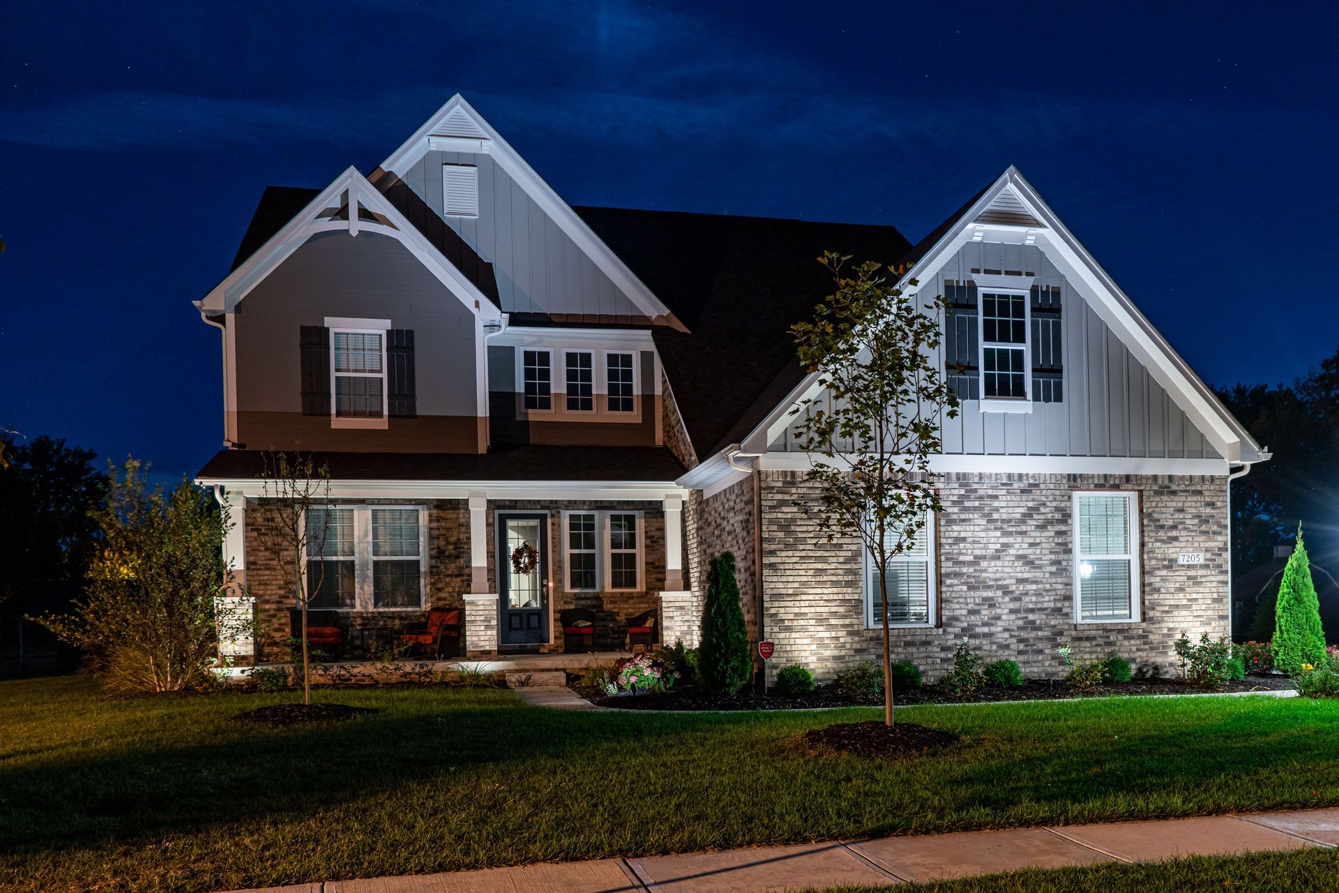 A two-story house at night with lights illuminating the exterior. Brick and siding with a front porch.