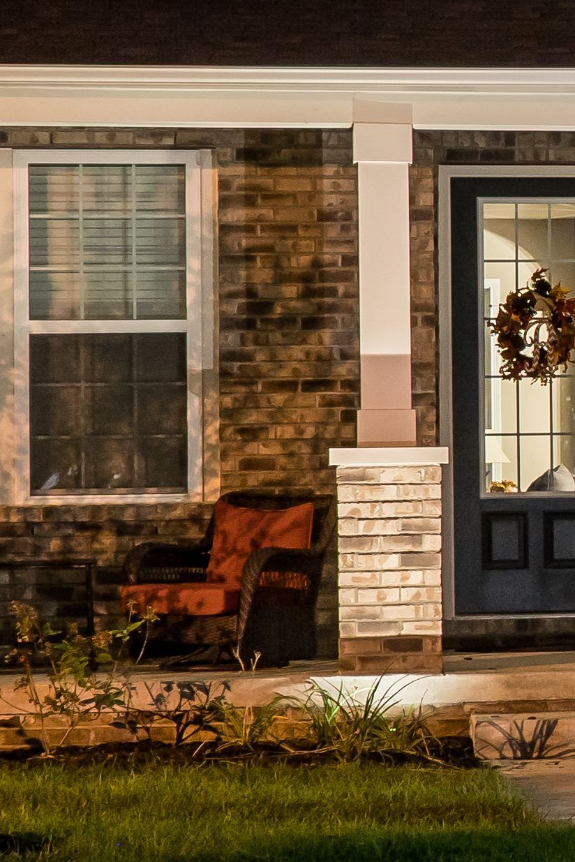 Exterior of a brick home with a front porch. A red armchair sits near a window, lit by nighttime lights.