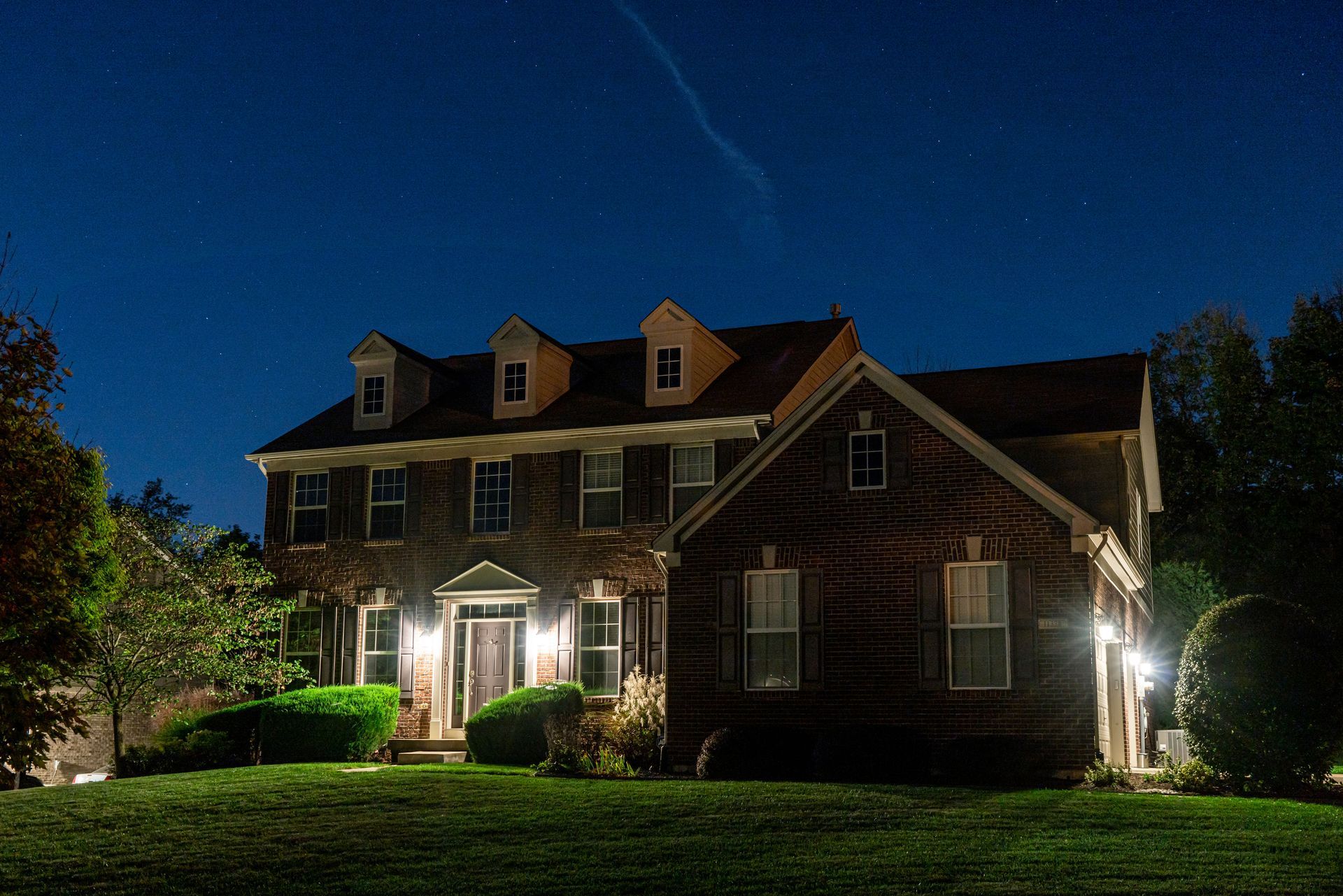 Two-story house at night; illuminated front door, windows, and lawn against a dark blue sky with stars.