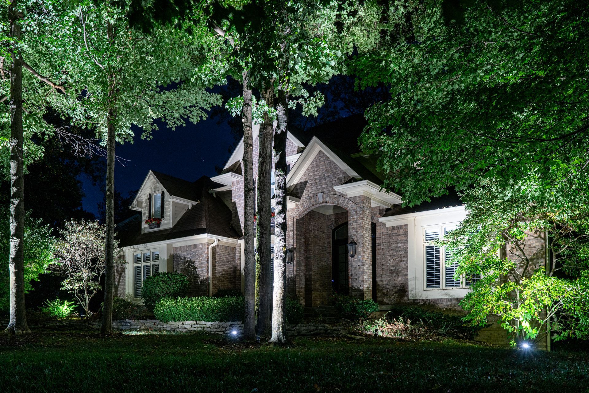 Night-lit brick house nestled among trees, a dark blue sky in the background.