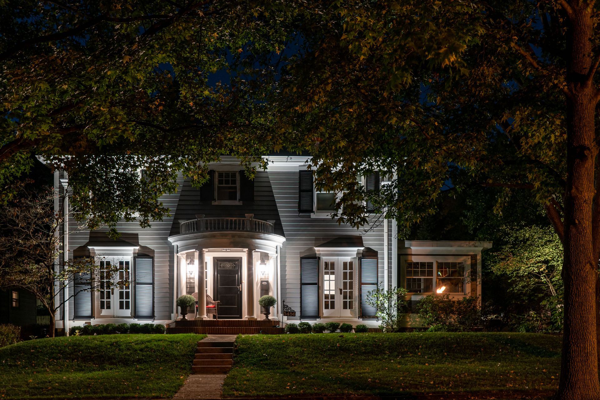 Two-story grey house at night, lit from within, with dark blue shutters and a tree in the foreground.