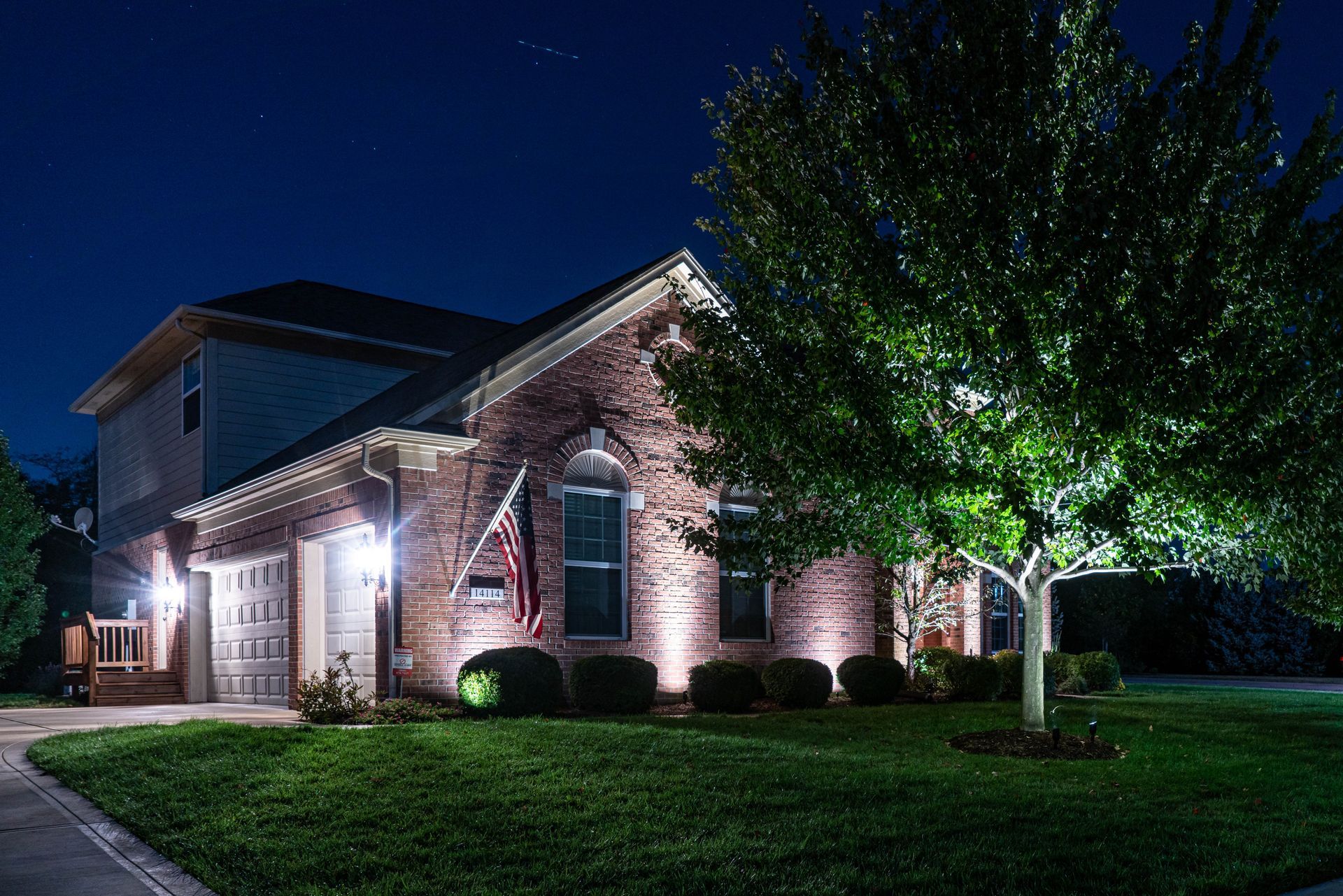 A brick house at night with landscape lighting illuminating the tree, lawn, and facade.