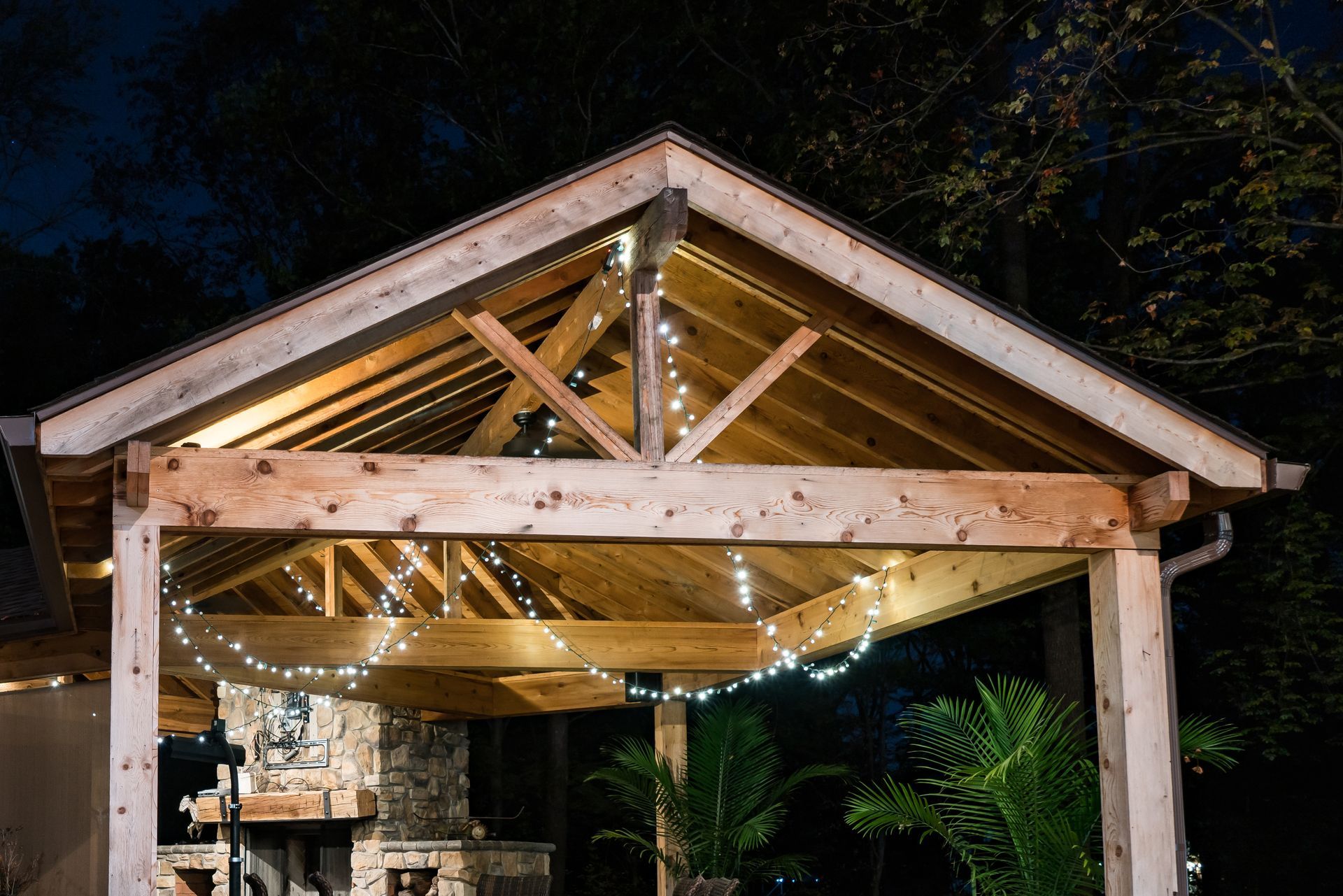 Wooden pergola with string lights, night setting, view of fireplace and greenery.