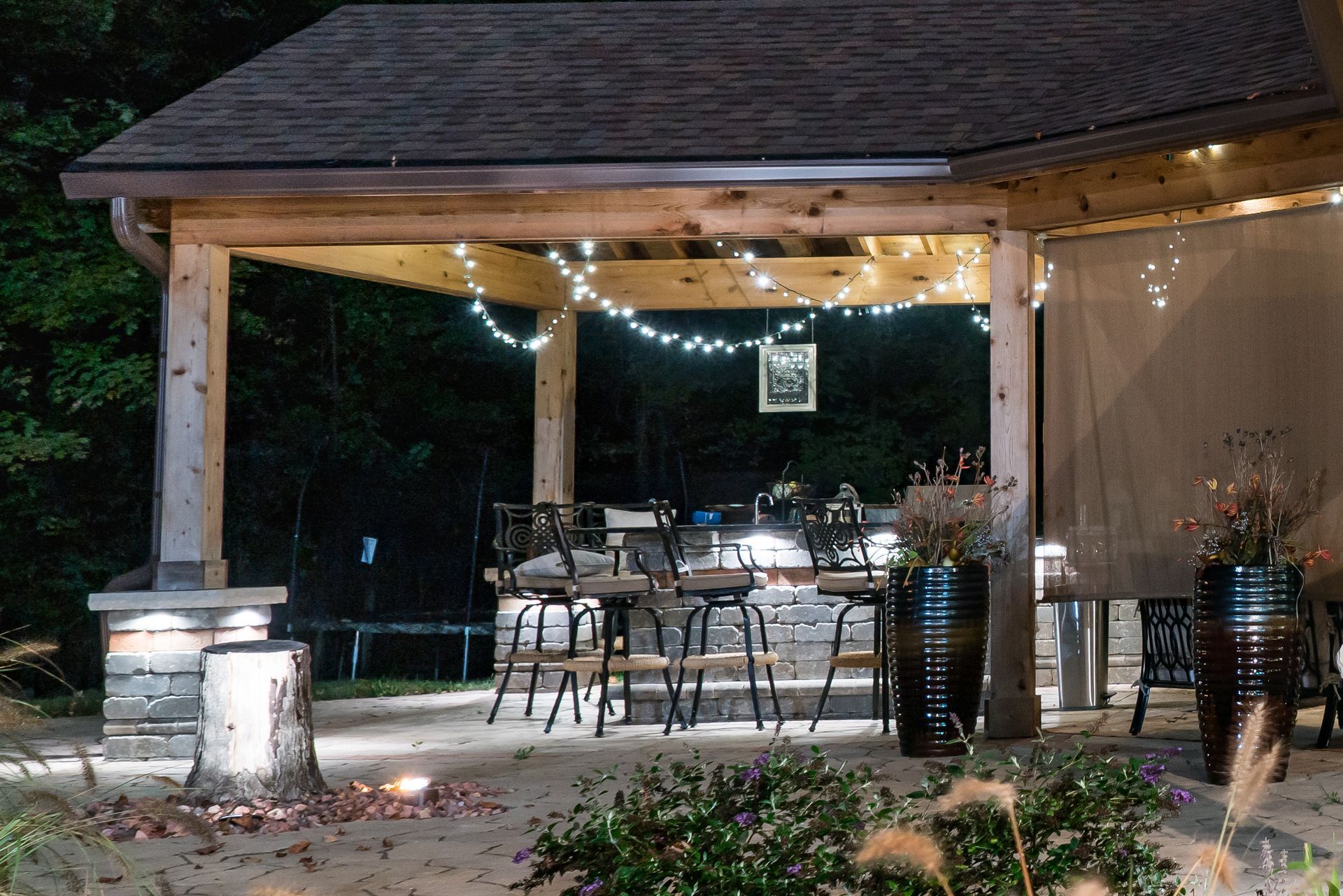 Outdoor patio with bar stools under a lit wooden canopy, with string lights and dark background.
