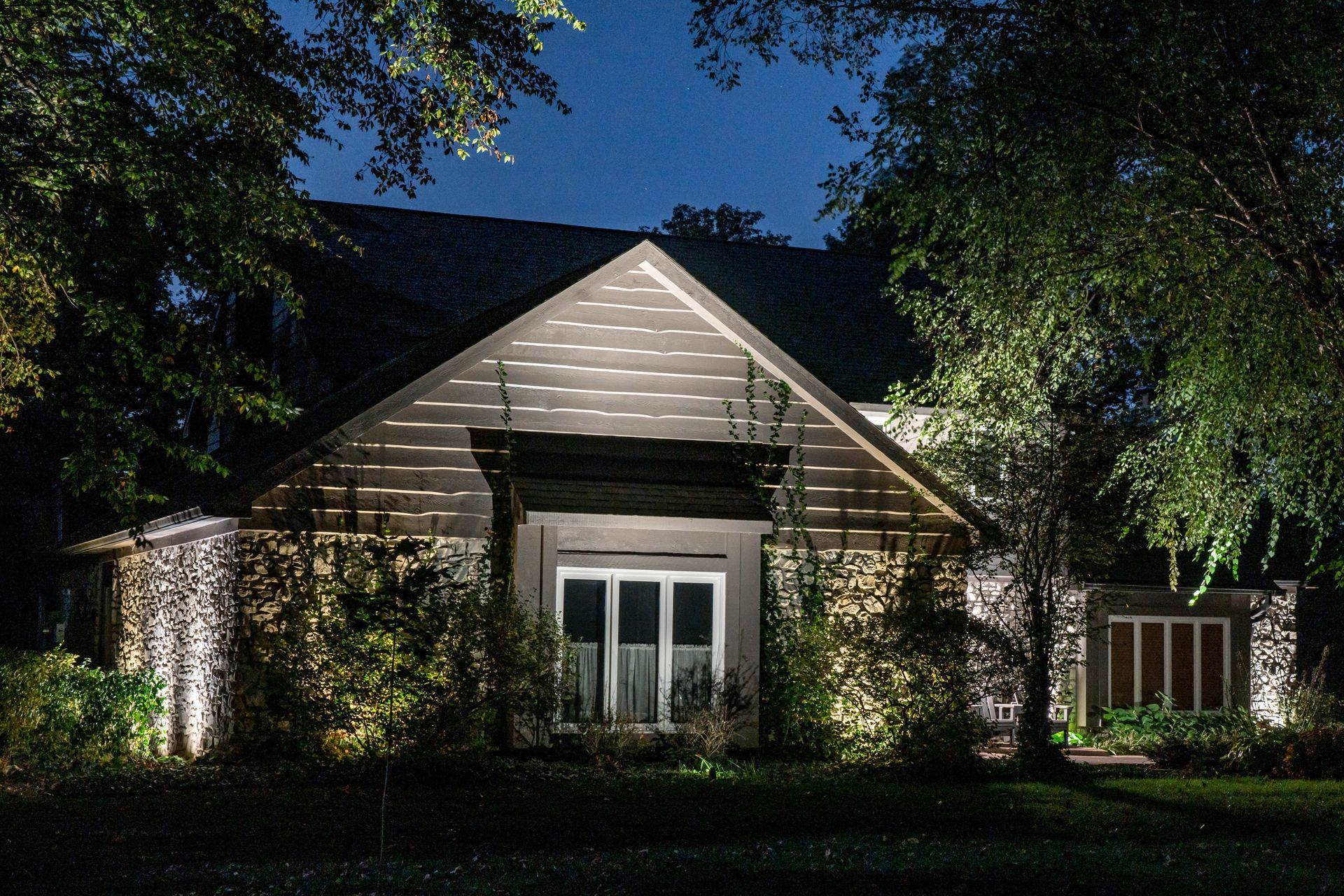House illuminated at night by landscape lighting. Stone exterior with front door and surrounding trees.