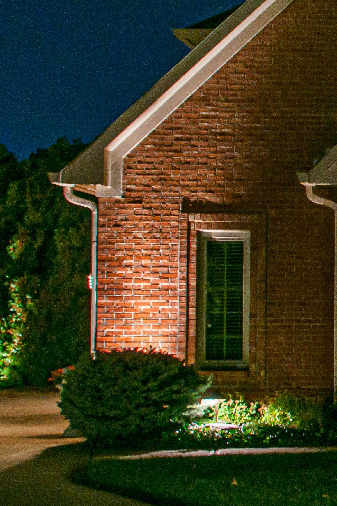 Brick house exterior illuminated at night, with a window and landscaping.