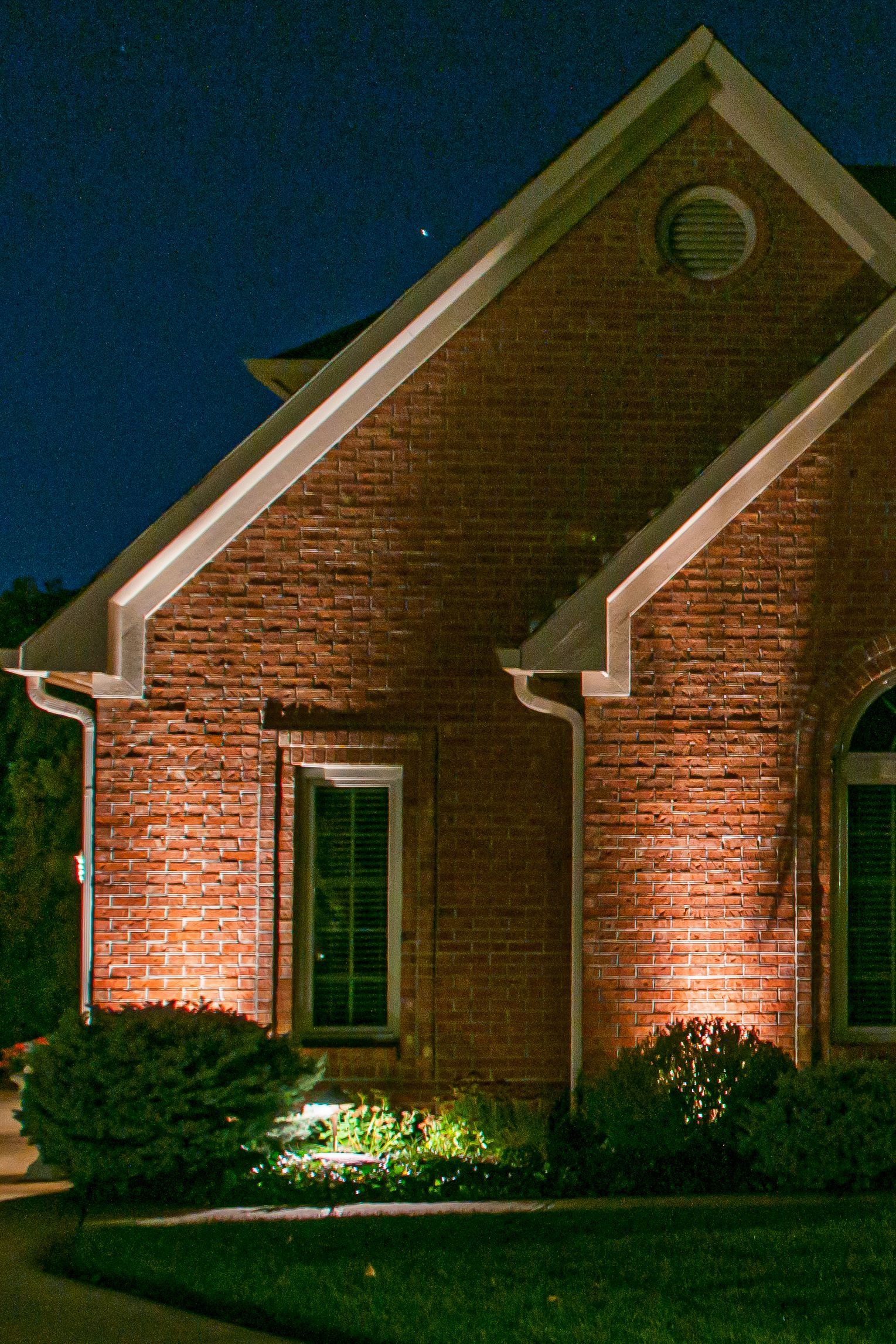 Brick house illuminated at night. Green bushes and grass in foreground, stars in the dark sky.