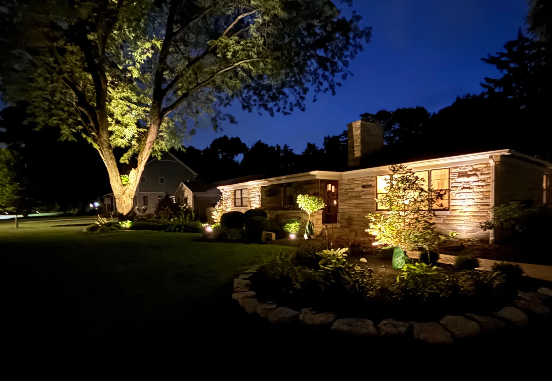 Night exterior house with landscape lighting illuminates tree, plants, and stone facade.