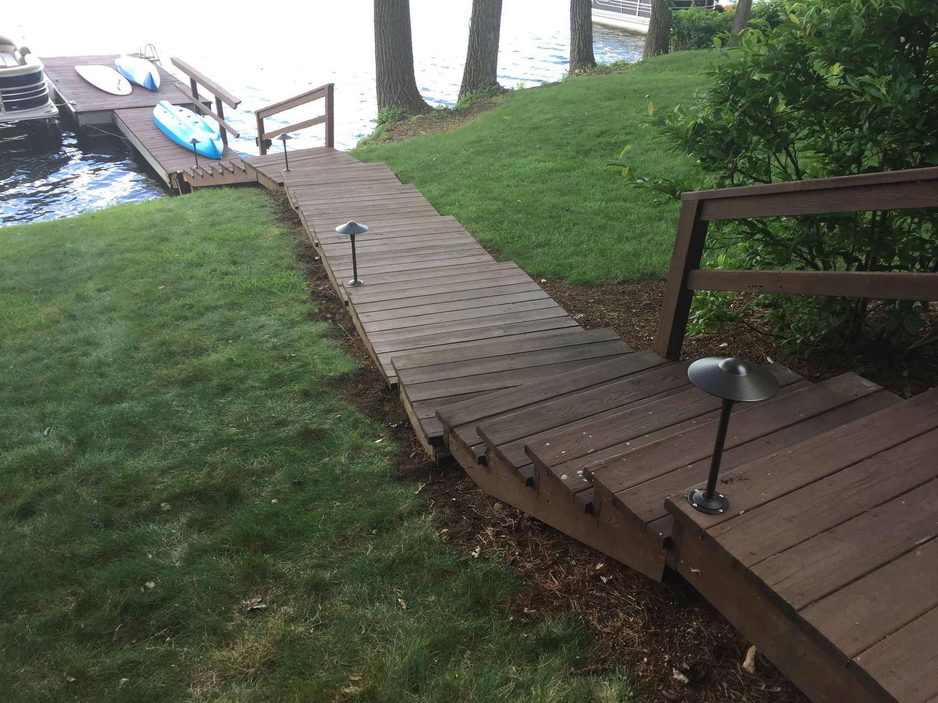 Wooden stairs leading down to a dock with kayaks on a lake, surrounded by grass and trees.
