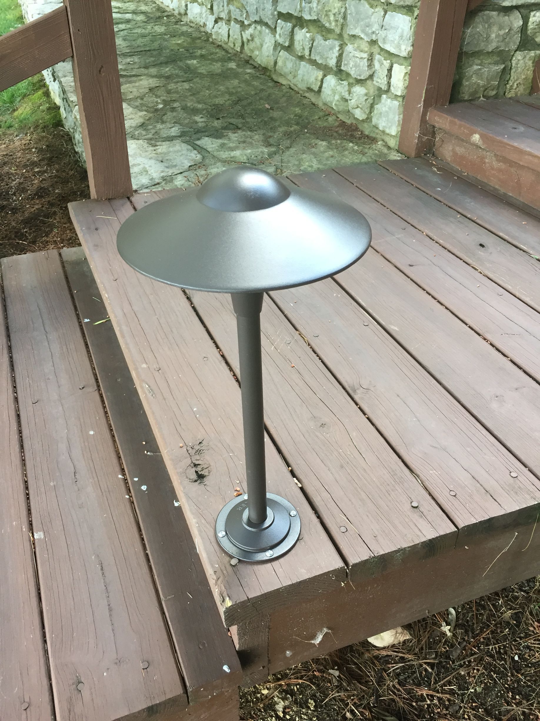 Dark gray outdoor light fixture on a wooden deck, with a stone wall in the background.