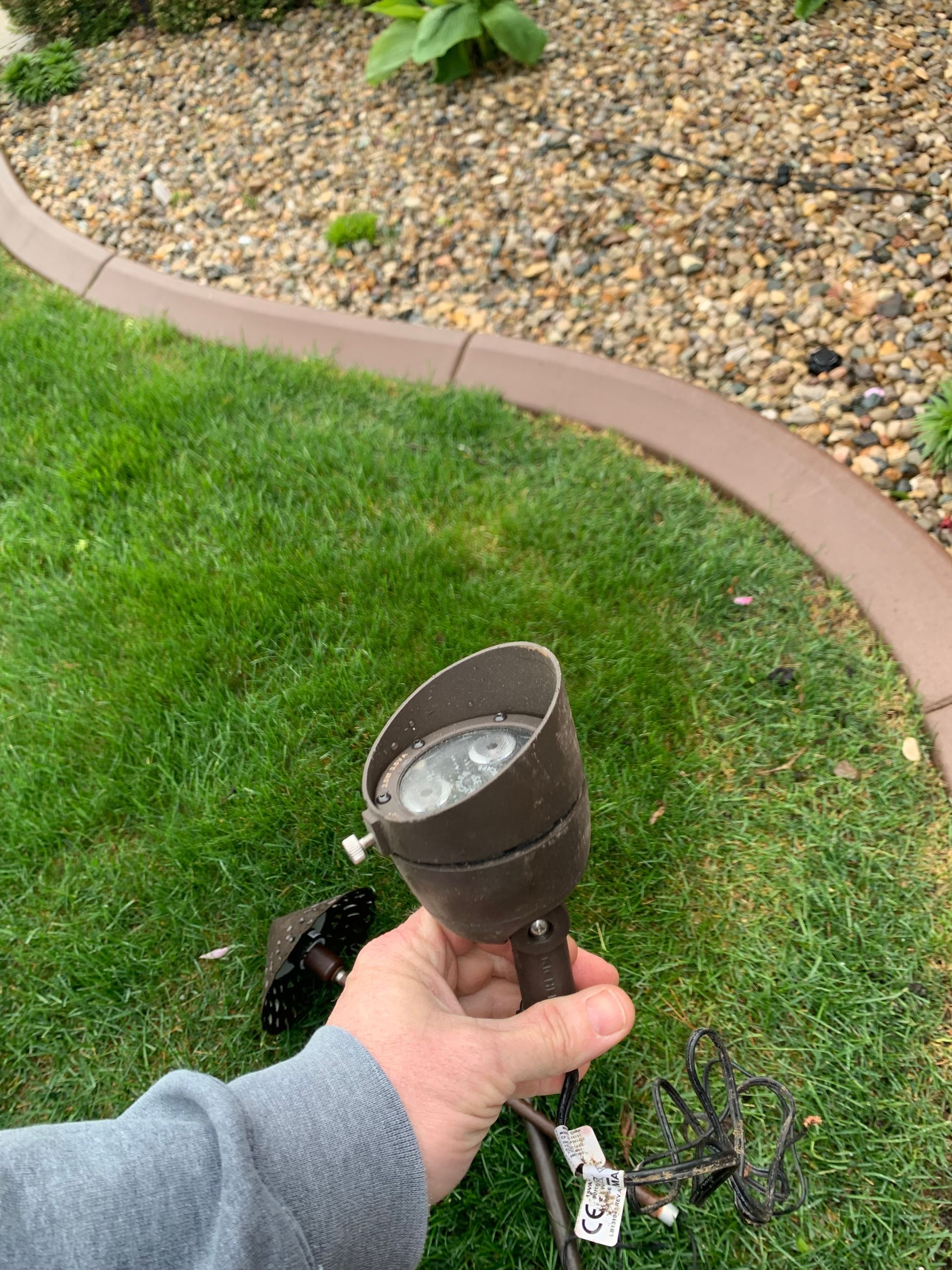 Hand holding a brown outdoor spotlight near grass and decorative stone edging.