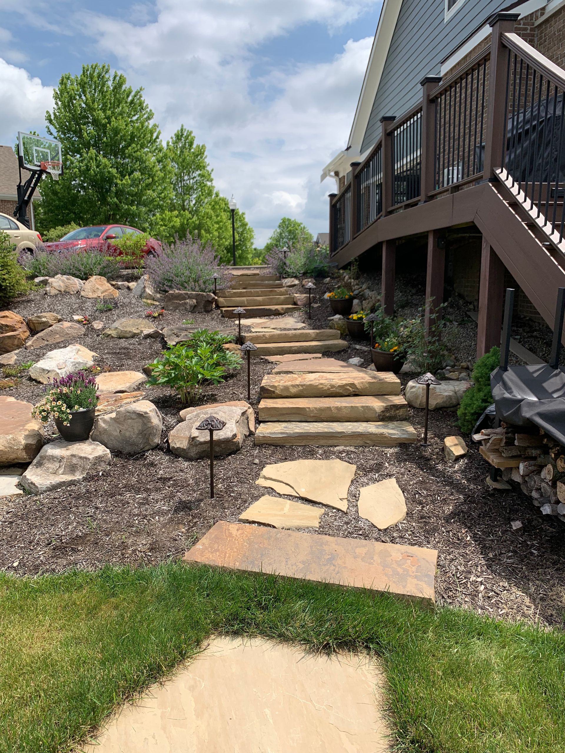 Stone steps and walkway ascend a landscaped backyard, next to a house with a wooden deck and railing.