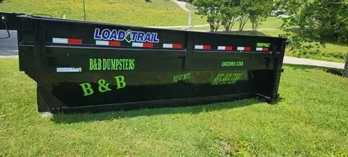 Black B&B dump trailer with colorful logos parked on a grassy lawn in front of trees