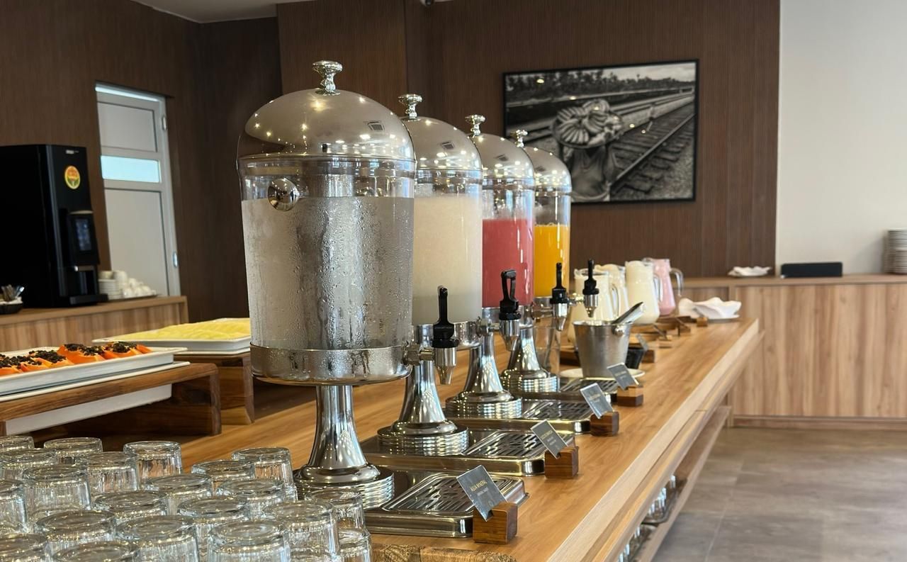 Mesa de buffet com dispensadores de bebidas, copos e alimentos, em uma sala de jantar.