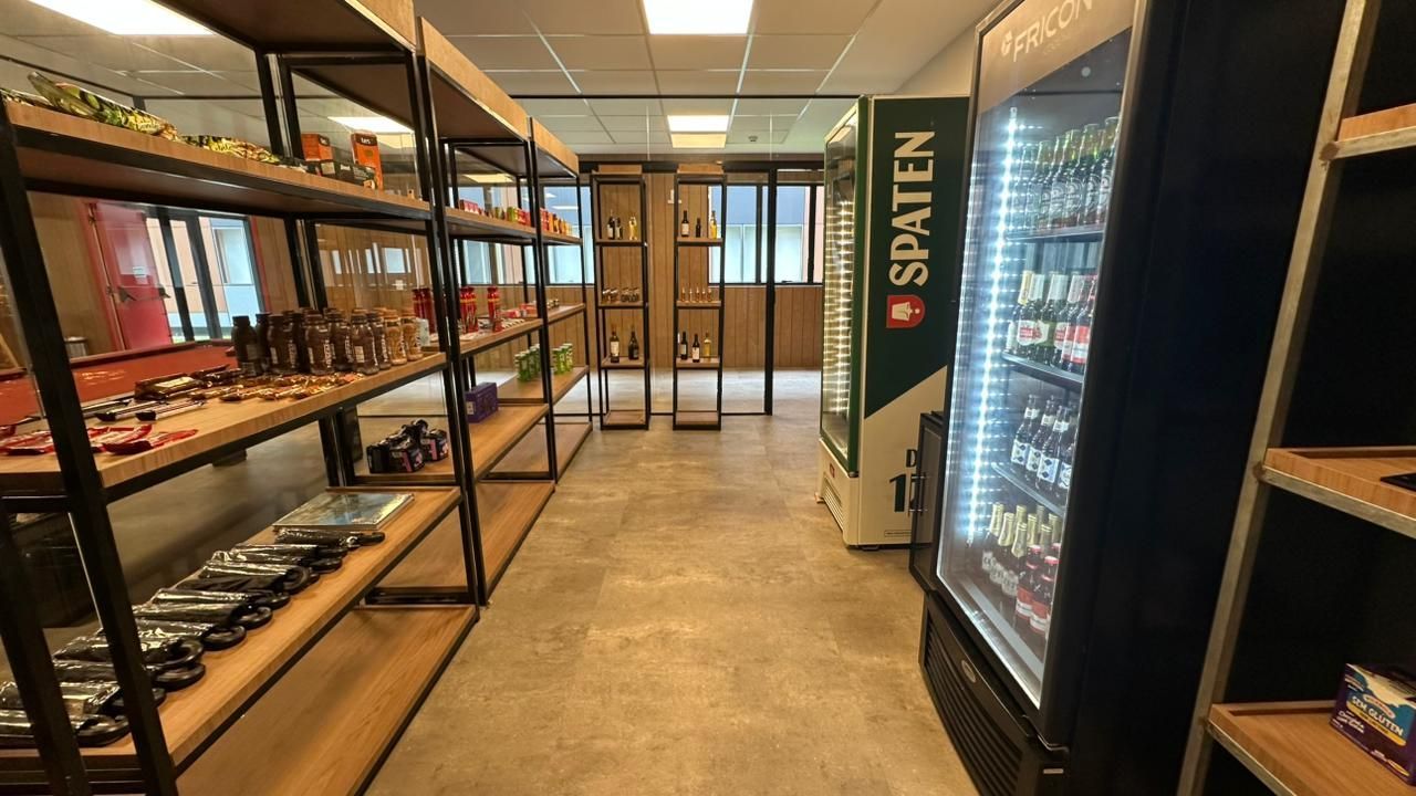 Prateleiras com lanches e bebidas, refrigerador Spaten e portas de vidro dentro de um escritório bem iluminado.
