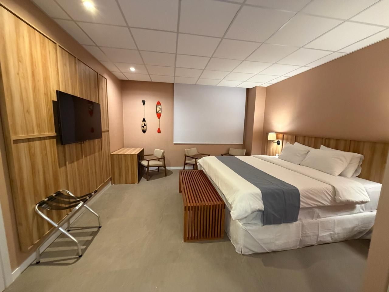 Quarto de hotel com cama, TV, escrivaninha e banco, com painéis de madeira e paredes cor-de-rosa.
