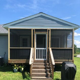 Screened-in porch with wooden steps and railing, blue siding, white door, and grassy yard on a sunny day.