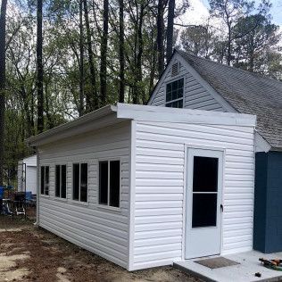 White-sided shed attached to a house with a door, windows, and a flat roof in a wooded area.