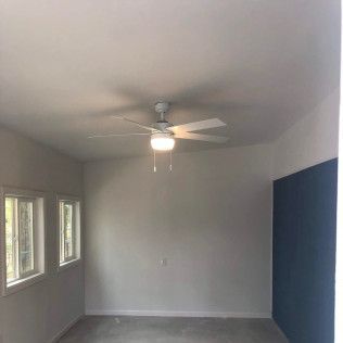 Empty room with a ceiling fan. White walls, windows on the left, and a dark blue wall on the right.