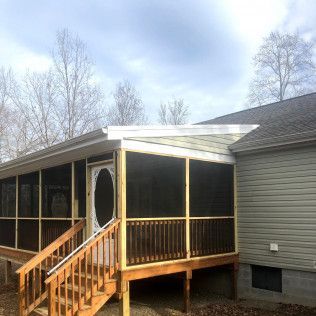 Screened porch with wooden deck, steps, and door attached to a house with gray siding under a cloudy sky.