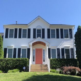 White two-story house with black shutters, pink front door, and green bushes against a blue sky.