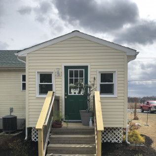 Small, cream-colored building with green door and stairs. Two windows flank the door. Cloudy sky in the background.