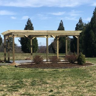 Wooden pergola in a grassy yard, surrounded by bushes and a circular stone border, with trees in the background.