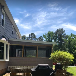 Screened-in porch on the back of a house with a grill and bench in the yard; bright blue sky.