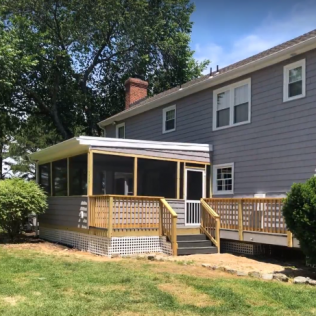 Gray house with screened porch and wooden deck, surrounded by green grass and trees.
