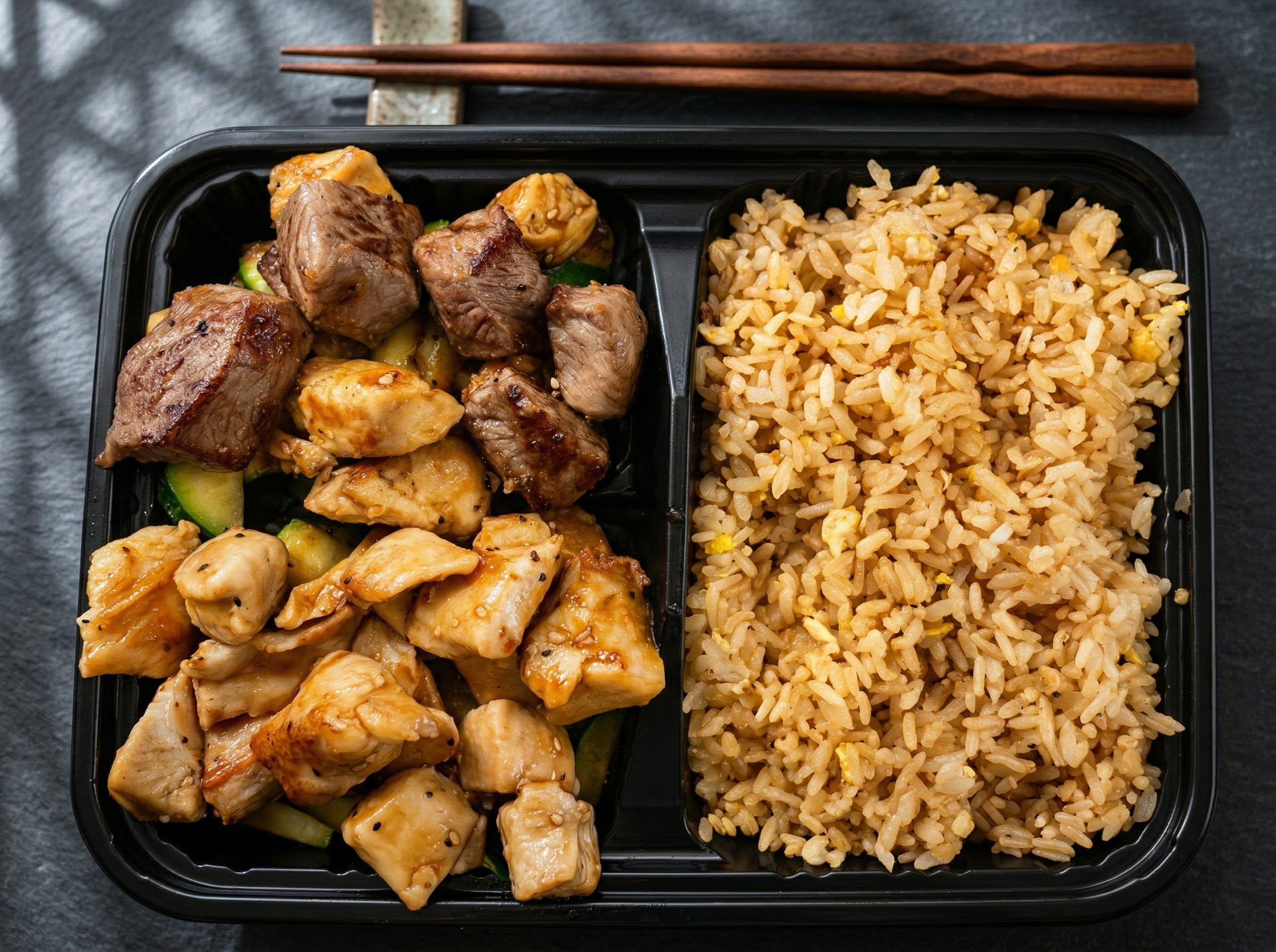 A takeout container filled with cubed steak, chicken pieces, and fried rice, placed next to a pair of wooden chopsticks.