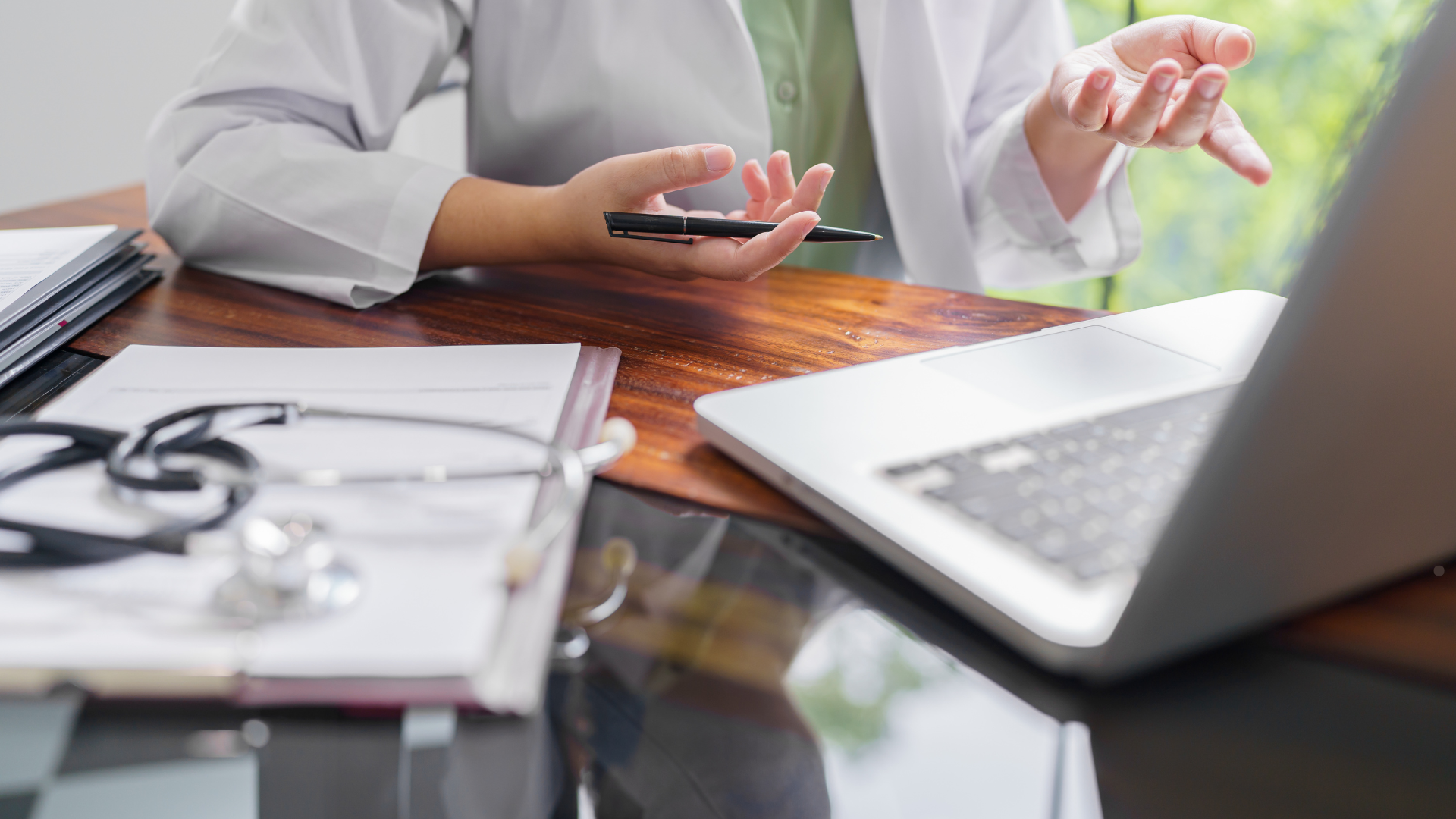 Healthcare provider discussing treatment during a telepsychiatry session, gesturing while using a laptop and holding a pen over clinical notes in a modern medical office.
