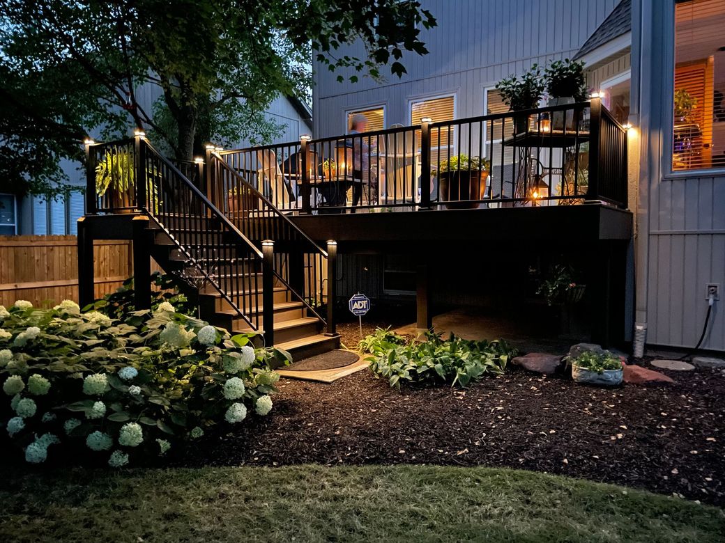 A lit-up wooden deck with stairs and black railings at dusk, surrounded by landscaping and house.
