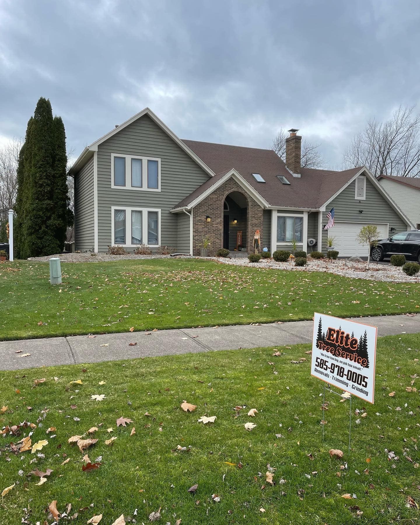 A two-story green house with a stone entryway, brown roof, and a lawn sign in front on a cloudy, autumnal day.