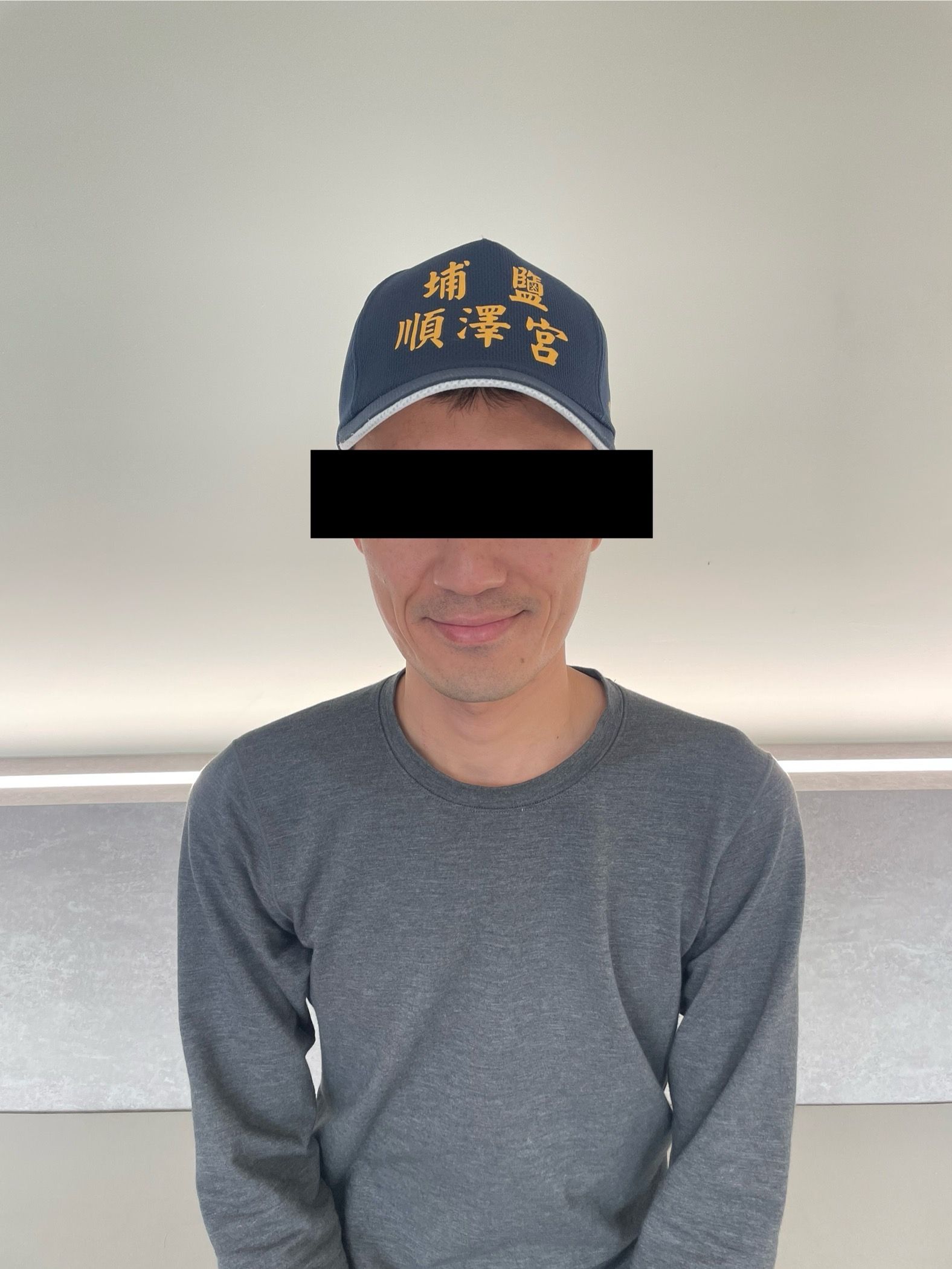 一名戴著印有黃色字樣的海軍藍帽子、身穿灰色襯衫的人，站在室內，靠著一面素色的牆。
