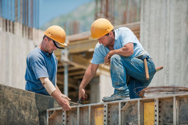 Dos obreros de la construcción con cascos amarillos miden encofrados de hormigón en una obra.