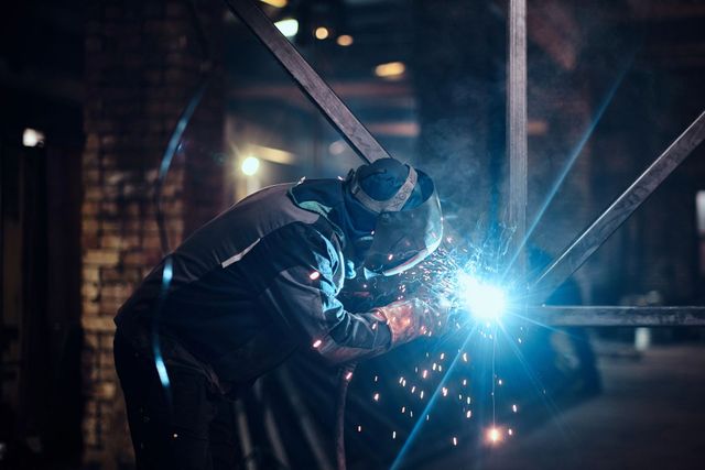 Un soldador con traje protector oscuro produce chispas de luz azul mientras suelda metal en un taller con poca luz.