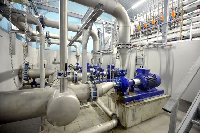 Sala de bombas industriales con motores azules, grandes tuberías plateadas y válvulas en una luminosa instalación mecánica.