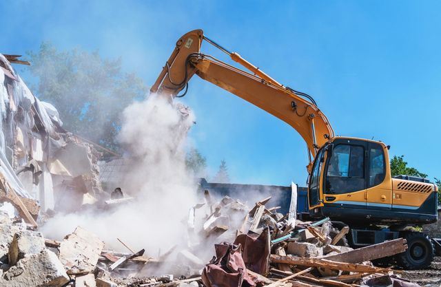 Una excavadora demuele un montón de escombros, levantando polvo bajo un cielo azul despejado.