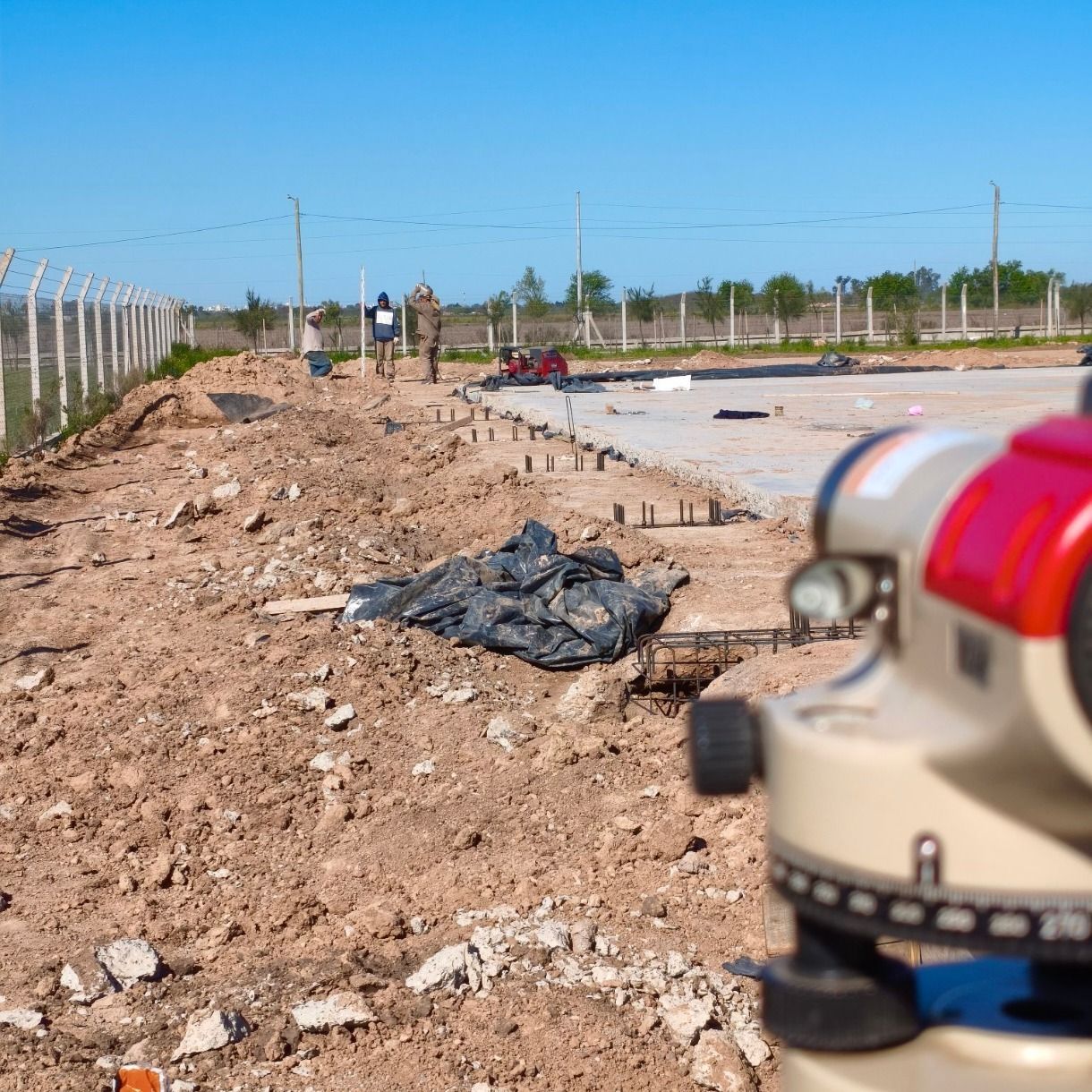 Obra en construcción con camino de tierra, valla y un instrumento de topografía borroso en primer plano.