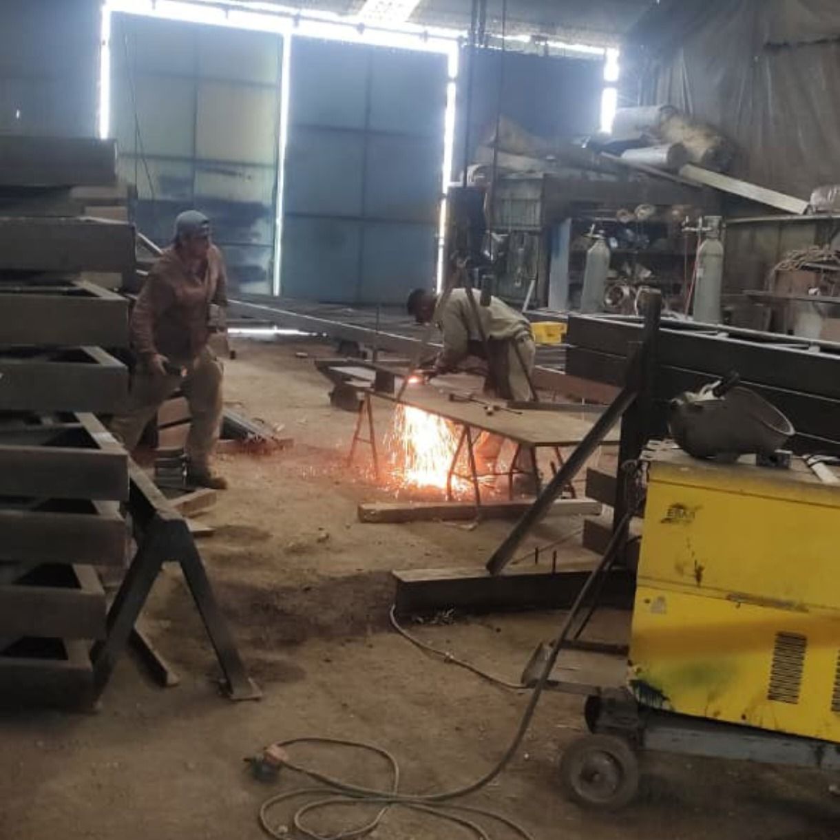 Trabajadores soldando metal en un taller desordenado, rodeados de chispas brillantes y maquinaria.