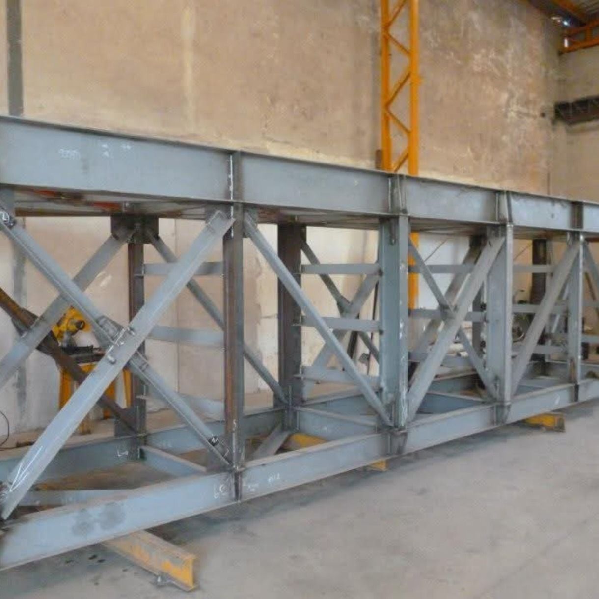Estructura de celosía de acero gris en el suelo de un almacén, con soportes amarillos al fondo.