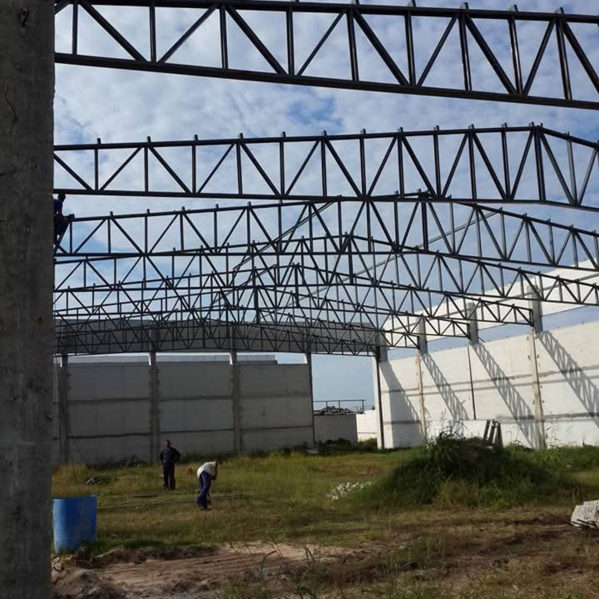 Estructura de acero de un almacén parcialmente construida, con trabajadores sobre un suelo de tierra cubierto de hierba.