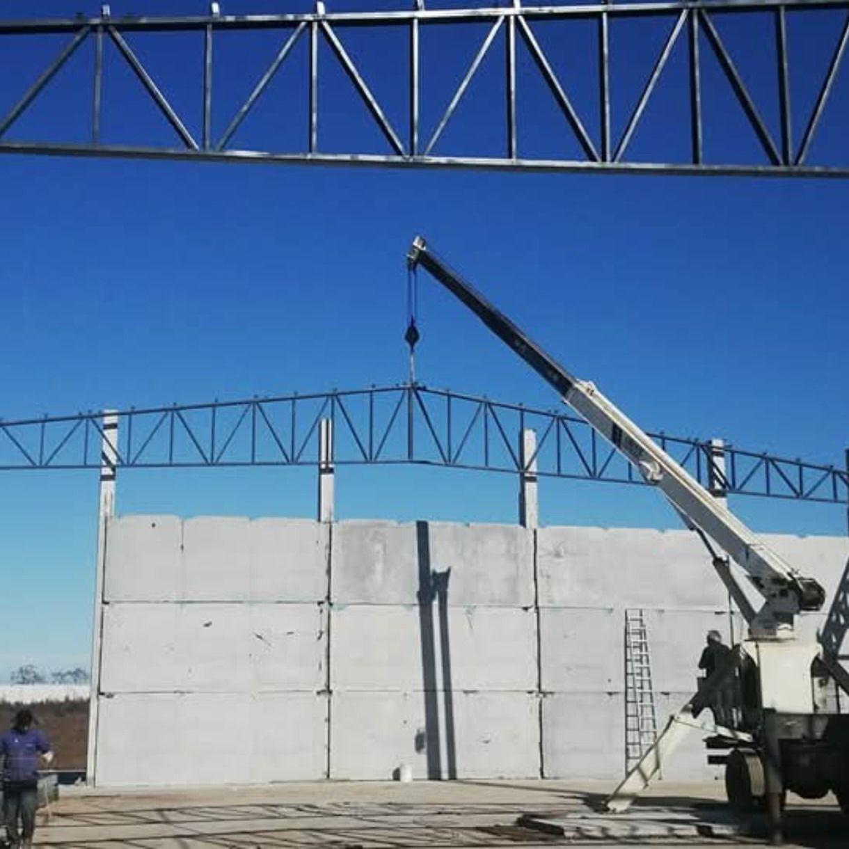 Obra en construcción con bloques de hormigón blanco y una grúa levantando una viga bajo un cielo azul.