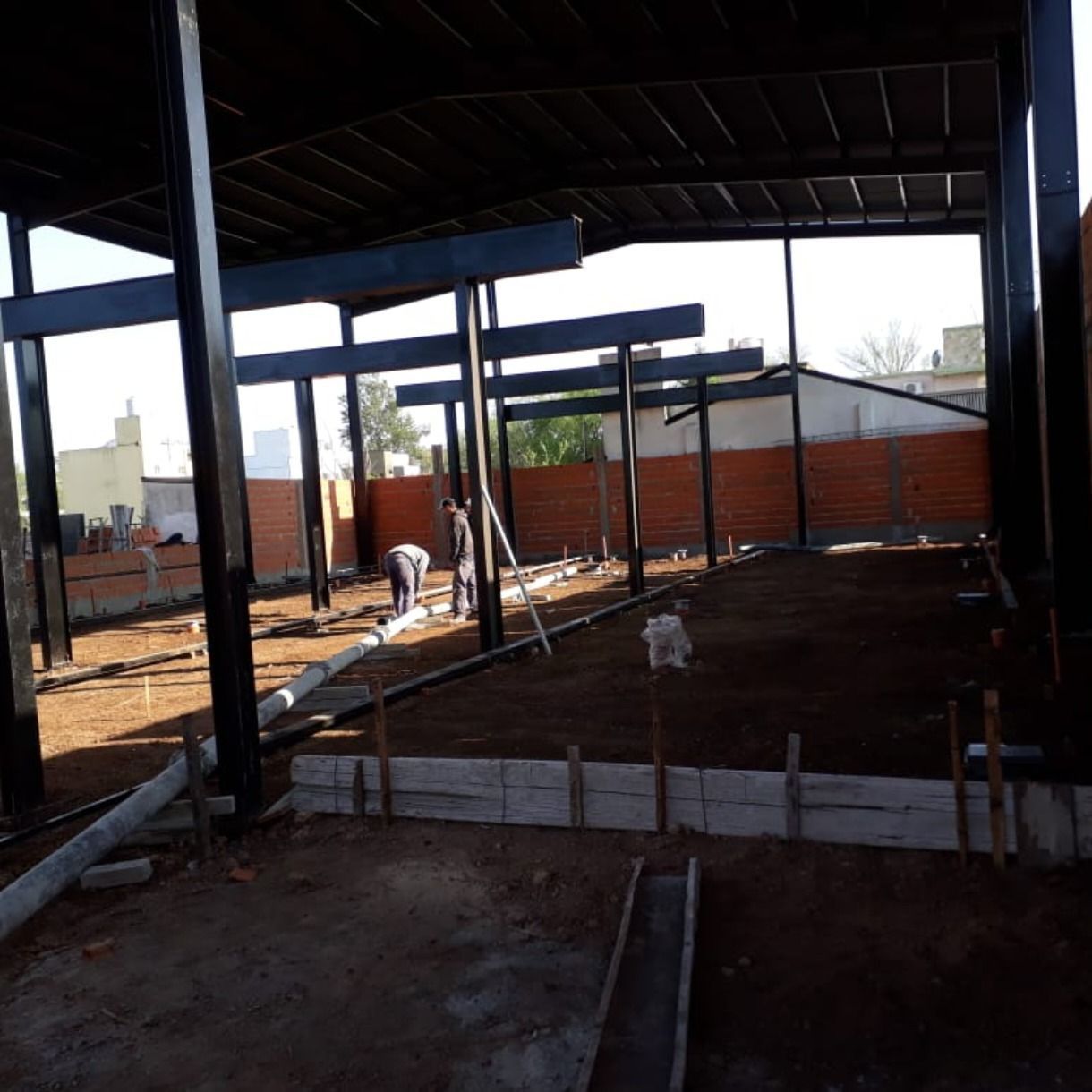 Interior de un edificio con estructura de acero en construcción, con trabajadores y paredes de ladrillo rojo al fondo.