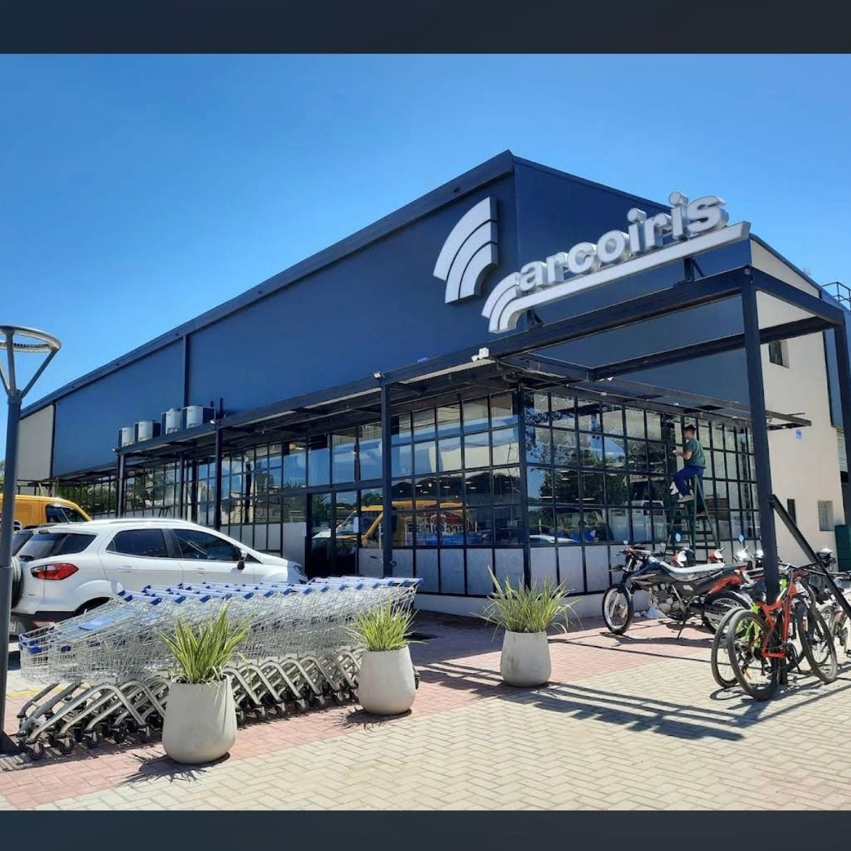 Exterior de una tienda Emco moderna con fachada de cristal, coches aparcados, bicicletas y carritos de la compra delante.