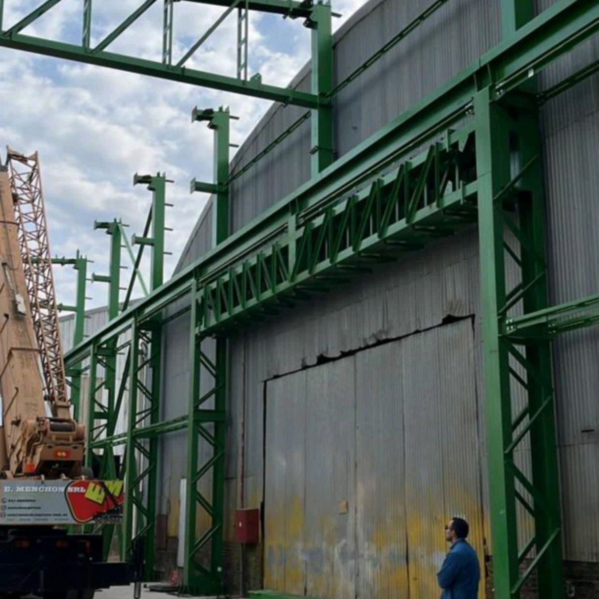 Edificio industrial verde con una grúa al lado y una persona de pie cerca de una gran puerta metálica.