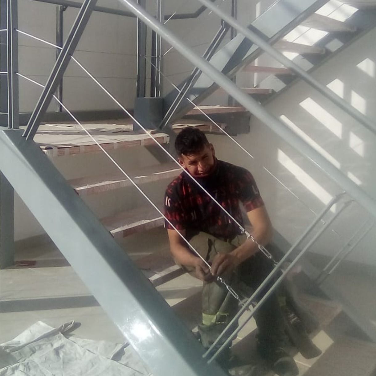 Trabajador utilizando una herramienta eléctrica en escaleras metálicas en un interior industrial luminoso.